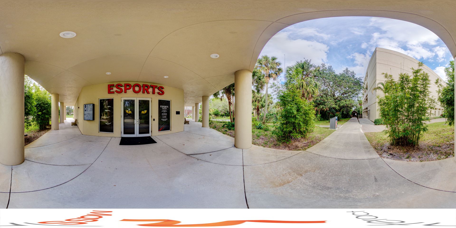 Panoramic view of the entrance to an Esports center, featuring double glass doors under a covered walkway, with lush greenery and palm trees surrounding the building
